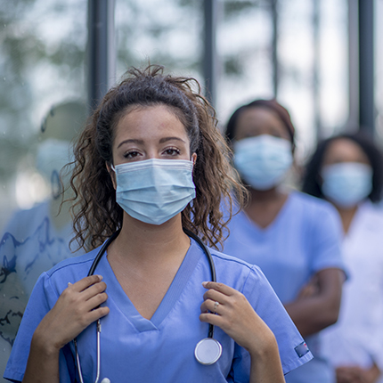 A group of nurses staring at the camera. They're in a line, with a woman in the front being visible. The two behind her are out of focus. They're wearing scrubs and face masks.