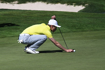Golfer wearing a yellow shirt, white baseball cap, and grey pants is preparing to putt the ball. 